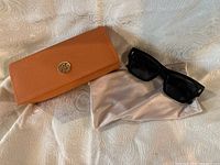 Photo showing black Tory Burch sunglasses resting on a beige protective pouch with an orange leather case beside them.