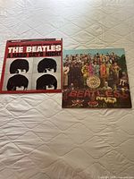 Front view of two Beatles vinyl LP albums side by side on a quilted white surface, showing covers of 'A Hard Day's Night' and 'Sgt. Pepper's Lonely Hearts Club Band'.