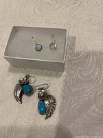 Two pairs of earrings on a beige textured fabric, one in small white box (round light blue stone earrings) and the other pair laid below with turquoise wing-shaped insets and silver flower motif.