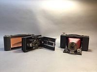 Two antique folding cameras displayed side by side, one open with red bellows visible, the other closed with a leatherette cover.