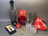 Eclectic barware set including a tall smoke gray Wedgwood decanter with stopper, red glass tilt bowl, clear rocks glasses, enamel dancing man bottle opener, Mr. Boston's Bartender Guide, and ceramics with tea towel.
