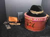 Top and side view of black Knox Hats felt hat with feather on band. Ernie label visible inside hat. Large round Knox Hats red box with emblem and leather strap.