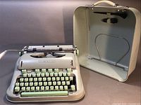 Full view of Hermes 3000 manual typewriter in pale green alongside its hard storage/carrying case with handle open showing interior metal attachments.