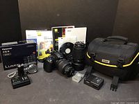 Full lot showing Nikon D5000 DSLR with attached Nikkor 18-55mm lens, Nikkor 55-200mm lens standing beside it, Nikon camera bag, batteries, chargers, software/DVDs, Panasonic Lumix ZS3 camera, battery and box.