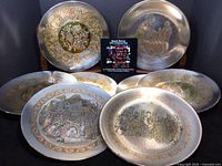 Eight silver plated collector plates arranged with visible detailed Christmas-themed Damascene artwork in gold and black on silver background, including figures and floral borders. Plates are visibly fragile.
