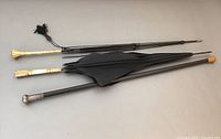 Full view of three items laid out: one black wooden cane with silver handle, one black parasol, and one parasol skeleton with gold and mother of pearl handles