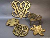 Five vintage brass trivets arranged on grey background showing worn brass patina, intricate designs including Williamsburg Queen Anne Cypher, Kings Arms crest, flower motif and heart shape with legs.
