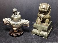 Front view of the lot showing the gray stone tripod vessel with Foo dog lid and dragon head handles, next to the green stone Temple Lion statue on pedestal.