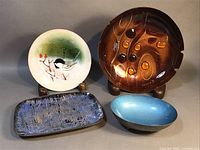 Four enamel on metal items displayed together: Norman Brummell Chickadee plate, Sascha Brastoff signed brown ash receiver/candy dish, small rectangular blue and black enamel plate, and Reed & Barton turquoise footed bowl.