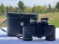 Binoculars displayed with black carrying case behind, showing textured black body and eyepieces.