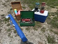 Full lot outdoors on grass and gravel showing all items: green Coleman camp stove, two Coleman lanterns (red and teal blue), wicker picnic basket with colorful plastic dishware, blue portable sun umbrella in carrying bag, and blue cooler box.