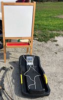 Children's wooden easel with whiteboard/chalkboard and red tray, and black torpedo snow sled with yellow handles and white pull rope, shown outside on gravel and grass.