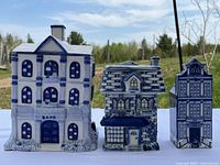 Front view of three handcrafted porcelain Delft collectible buildings: a bank, a cottage, and a tall narrow building, all painted in classic blue and white.