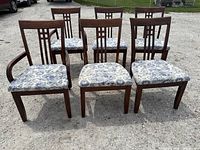 Photo showing six of the seven wooden dining chairs arranged outdoors on gravel surface, including side chairs and captain's chairs with upholstered seats having a grey-beige floral pattern.