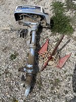 Full view of Evinrude 2 HP trolling motor lying on gravel with two red boat anchors and rope beside it.