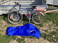 Side view of the entire blue Fisher Advance mountain bike with red rear rack and two red bottle holders on frame, alongside the blue bike cover laid on the ground.