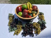 Wooden bowl filled with various artificial fruits and vegetables, surrounded by bunches of artificial grapes on a white cloth.
