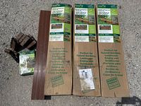 Three unopened rectangular kits of garden and playground border edging kits with signage describing them as perfect for raised gardens, landscape edging, and sandboxes, each 16 feet long, placed on pavement alongside additional used pieces of the material.