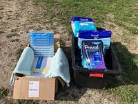 Box containing Batrik Saveface isolation face shields and light blue mattress protectors and gowns, next to black bin with multiple Prevail Overnight incontinence product packages.