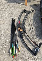 Photo showing two junior compound bows and a set of arrows on pavement surface, bows have pulley systems and curved limbs, arrows have various colored fletching.