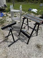 Photo of a multi-position aluminum ladder, foldable black Workmate bench with measuring grid surface, and adjustable black metal saw horse on gravel outdoors.