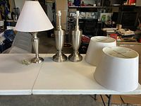 Three lamps displayed on a folding table with their respective fabric lamp shades beside them. Two larger matching lamps show urn shape bases and one smaller lamp with a simpler design. Shades are white or light colored with some visible staining.