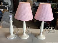 Three white bedside lamps, two with pink checkered fabric lampshades and one without shade, placed on table in warehouse setting.