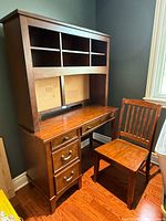 Desk with attached upper cabinet and the wooden chair placed adjacent. Desk has three drawers with metal handles and open shelves on the upper cabinet.