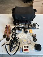 Photo of Minolta XG-1 camera with 50mm lens, camera bag, camera strap, lens caps, and cleaning supplies spread on white table.