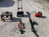 Wide shot of assorted outdoor tools including Vermont American Router/Saber Saw Table, Poulan Pro trimmer, Noma snow thrower, and red air compressor tank on gravel ground.