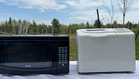 RCA black microwave and white Black & Decker bread maker side by side on white surface outdoors with trees in background.