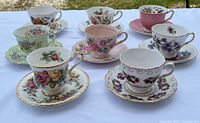 Overview of 8 assorted vintage tea cups arranged with their matching saucers on white backdrop under daylight, showing floral and pastel designs with gold accents.
