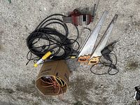 Photo showing the full lot including vise, two saws, electric drill, extension cord, and cardboard box with wires on concrete ground