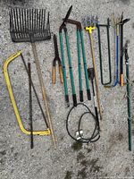 Photo showing general overview of all outdoor yard tools including rakes, shears, loppers, cultivators, pruners, saw, and cable lock arranged on gravel surface.