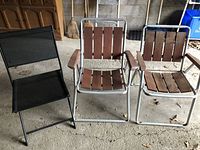 Three folding lawn chairs arranged side by side on a cement floor in a garage or similar space, showing two with cedar wood slats and one with navy vinyl