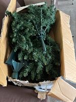Top-down view of partially assembled green artificial Christmas tree branches and green plastic stand inside a damaged cardboard box.
