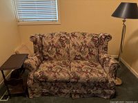 Floral loveseat with side table and floor lamp positioned around it in a room corner with window blinds.