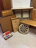Wide shot showing the cribbage table, dartboard, dart set, folding card table, and four metal folding chairs against a wall.