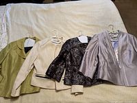 Four ladies formal jackets in lime, cream, black floral, and lilac colors arranged on hangers on beige textile background.