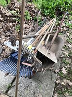 Photo of rusty garden tools piled in a metal wheelbarrow, including shovels, rakes, broom, hoe, pitchfork and rotary cultivator with wooden handles.