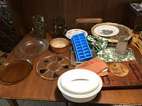 Wide view of the kitchen items arranged on a wooden table including glass pitchers, serving dishes, utensils, placemats, and ice cube tray