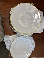 Eight white porcelain dinner plates stacked, each with a gold trim edge, on a table with packing materials visible.
