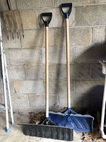 Two large snow shovels with wooden handles and plastic heads, one black and one blue, standing against a concrete wall.