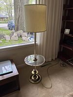 Full view of floor lamp next to window showing cream fabric shade, brass pole, glass tabletop and brass base.
