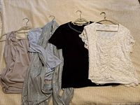 Four women's tops laid flat on a light beige surface. Tops include beige silky ruched blouse, silver sequined wrap top, black knit top with bead details, and white lace short-sleeve top.