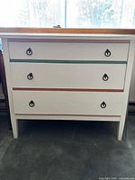Front view of the wooden dresser showing the three drawers with colored stripe accents in teal, terracotta, and pink and black metal ring pull handles