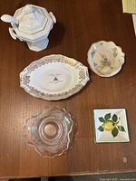 Top view of vintage ceramic gravy boat with ladle, assorted dishes including Royal Stanford Bone China dish, Red Cliff Iron Stone tray, small floral dish, clear pressed glass dish, and lemon motif square dish all arranged on wooden table.