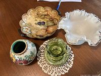 Four vintage decorative items displayed on wooden table: porcelain bowl with floral and fruit design, green glass dish with ruffled edge, round floral porcelain vase with repaired crack, and textured green glass small bowl.