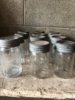 Set of twelve clear glass pint Crown jars with metal zinc screw rings and glass lids, placed on a wooden surface against concrete wall with visible dirt and wear. View from front and side showing embossing and general condition.