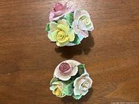 Two small porcelain flower bouquet figurines with pink, yellow, and cream roses, and green leaves on round white bases shown on wooden surface.